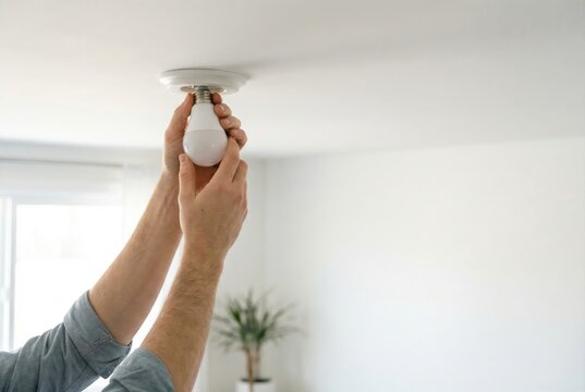 A person's hands screw a new energy-efficient LED light bulb into a white ceiling light fixture.