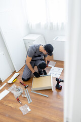 Worker assembles furniture in a bright room with tools on the floor and plans scattered around