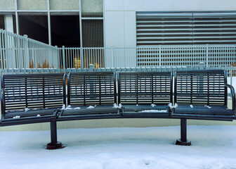 Snow covers a curved metal bench near a building in winter with a white background and a fence in...