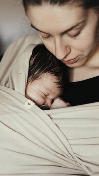 Close up of a mother holding her sleeping newborn baby in a beige fabric wrap carrier. Vertical shot of a woman with an infant in a sling indoors