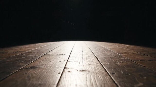 A dramatic low angle view captures the texture of aged wooden floorboards illuminated by a single bright spotlight against a deep black background creating anticipation.