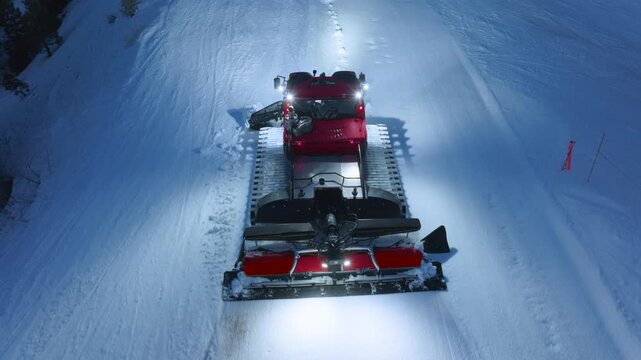 VARIANT 1 DESCRIPTION: Close up drone shot of a snow groomer at night leveling fresh snow on a ski slope. Bright headlights cut through the dark at a winter mountain resort.