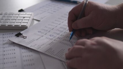 A person is focused on writing notes on a piece of paper while seated at a desk. A calculator and various documents are nearby on the table during daylight hours.