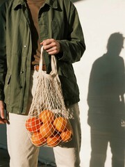 Person holding a net bag of oranges, casual lifestyle depiction, promoting sustainable shopping choices.