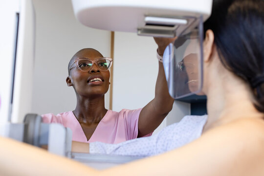 African American tech in pink scrubs adjusting mammogram paddle, female patient standing in gown