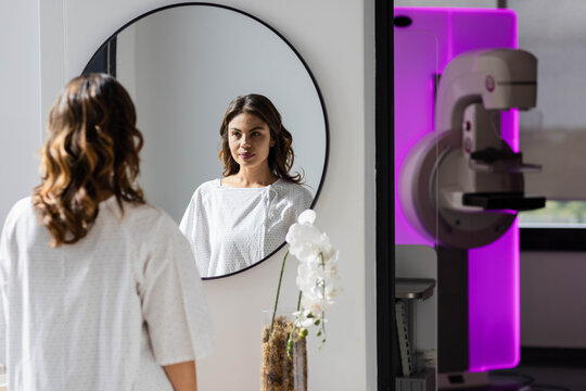Woman in her thirties standing in medical gown, looking into round mirror near mammography machine