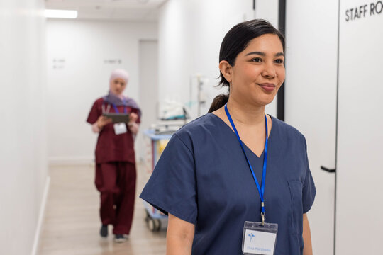 Diverse female staff walking hospital corridor, navy scrubs nurse showing ID, colleague with tablet