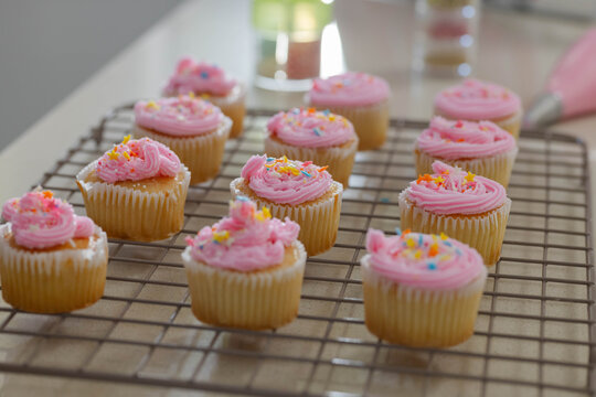 Vanilla cupcakes are cooling on metal rack on beige countertop with pink frosting and sprinkles