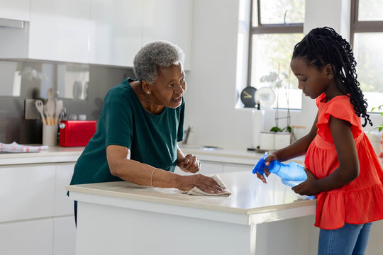 African American senior woman and child girl wiping kitchen island with blue spray bottle and cloth