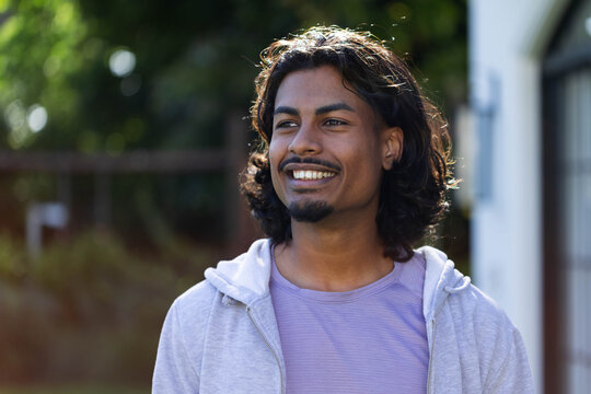 Indian man standing smiling while looking right in backyard near wooden fence wearing gray hoodie