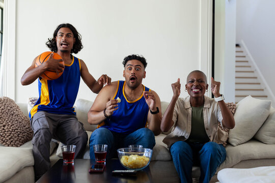 Three friends cheering and reacting on light sofa at home wearing blue jerseys holding basketball