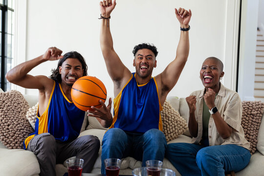 African American friends cheering, wearing blue yellow jerseys, holding basketball on sofa at home