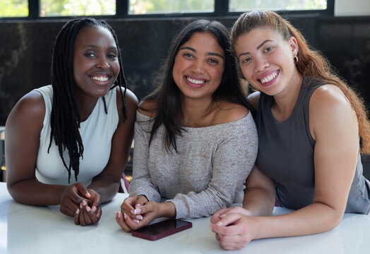 Adult female friends sitting close at countertop, leaning forward smiling by window with smartphone