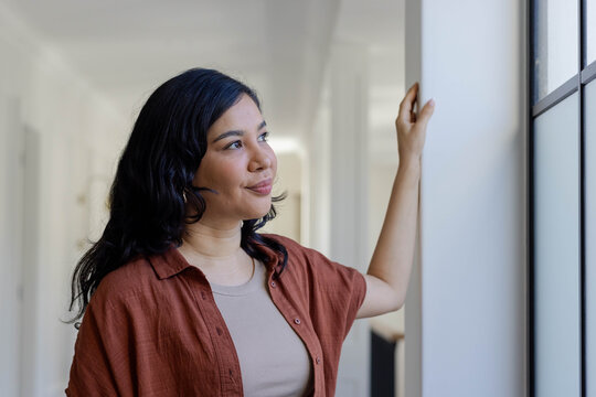 Female leaning on column in corridor, rust shirt, hoop earring, gazing at frosted window with doors