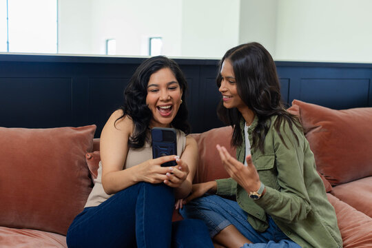 Diverse female friends laughing, leaning in and sharing smartphone on rust sofa in living room