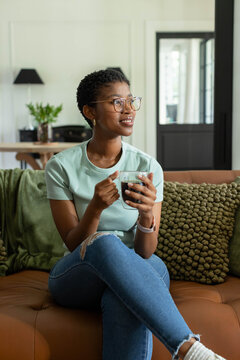 Adult African woman relaxing on sofa at home wearing mint top, ripped jeans, holding clear mug