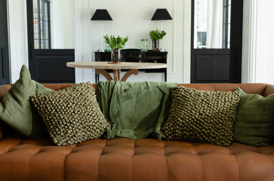 Brown tufted leather sofa is sitting centered with three green pillows and blanket in living room