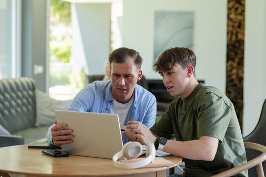 Father and teenage son using silver laptop at home, leaning, son wearing olive tee with smartwatch