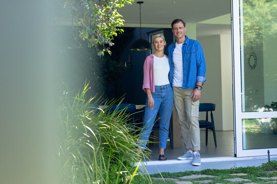 Couple standing in doorway of home wearing casual clothes, framed by sliding glass door and grasses
