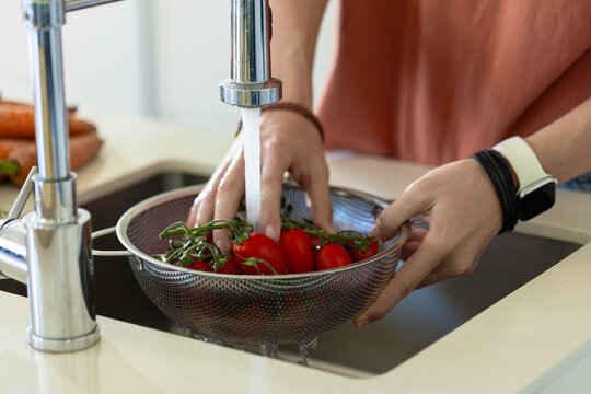Hands rinsing red vine tomatoes in steel mesh colander under running chrome faucet, carrots nearby
