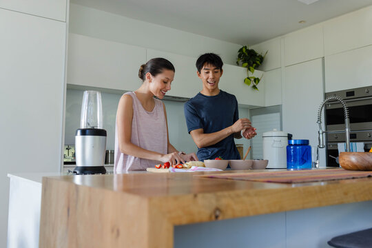 Couple slicing fruit on cutting board and stirring bowl at wooden kitchen island with blender