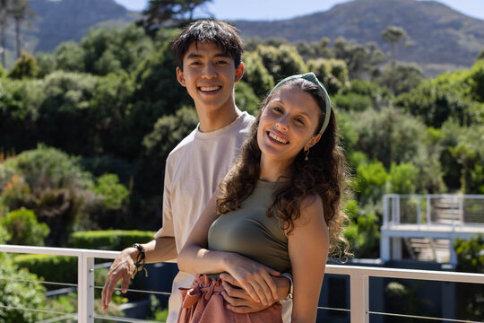 Asian man and African American woman standing on balcony railing, holding waist, smiling at camera