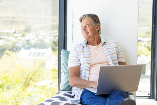 Senior man sitting on window seat using laptop, wearing striped shirt, patterned throw and cushion