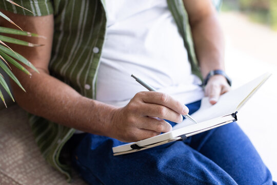 Silver pen is writing in open hardcover notebook on lap, green shirt and plant leaves visible