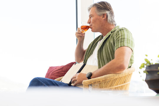 Mature male sipping amber drink at window holding tablet on lap in green shirt, copy space