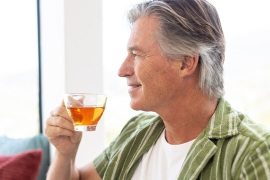 Senior man wearing green striped shirt sitting on sofa near window holding glass cup amber tea