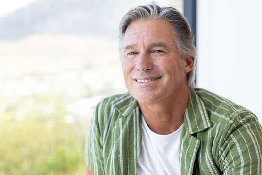 Senior man sitting beside large window, smiling gently, wearing green striped shirt, natural light