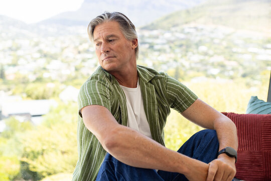 Senior man sitting at home by large window, wearing green shirt and watch, red, teal cushions