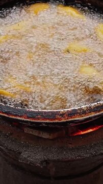 Close up view of deep frying plantain in old metal saucepan with dark or black burnt spots. Frying plantain in hot oil on coal pot. Orange charcoal flame inside coal pot.