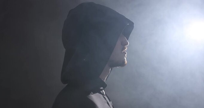 Man in hood turning around on dark background with light and smoke, camera moving up