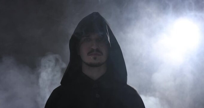 Man in hood turning around on dark background with light and smoke