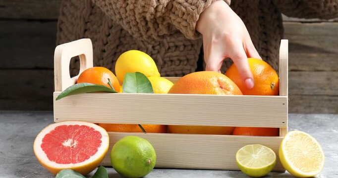 Woman putting fresh citrus fruits into wooden crate at grey table, closeup
