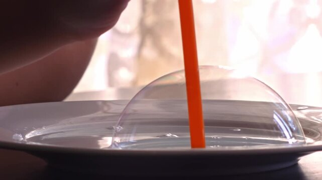 Close-up shot of a science experiment as a straw is used to blow air into soapy liquid, forming a translucent bubble on the surface.