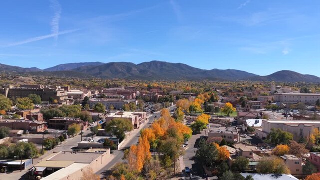 A sweeping drone perspective captures the historic Santa Fe skyline during peak autumn. Vibrant yellow trees weave through adobe architecture, leading to the rugged blue mountain range on the horizon.