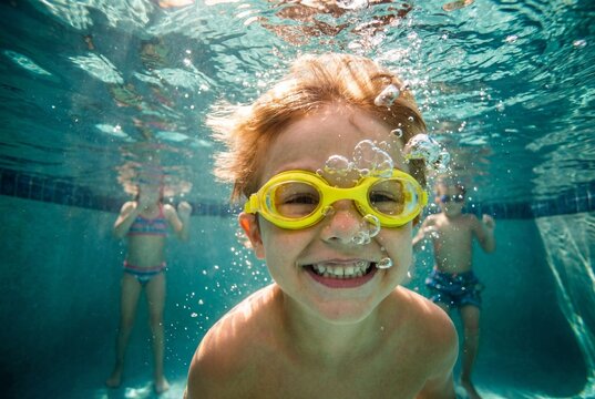 Joyful young boy wearing yellow goggles and smiling broadly underwater in a swimming pool with bubbles everywhere