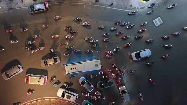 Aerial top-down night view of dense traffic in Ho Chi Minh City. Motorbikes and cars navigate busy urban intersection, showcasing chaotic flow and nightlife in Vietnam.