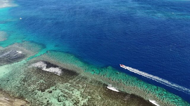 Drone orbit around Eua Island, Kingdom of Tonga, sweeping above coral reefs as a small speedboat cuts across turquoise water, leaving a bright foamy trail through the untouched Pacific paradise.