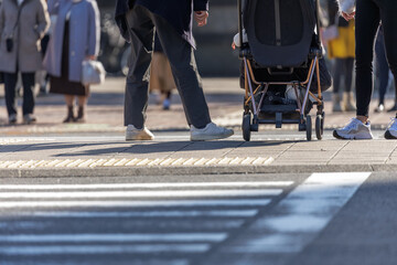 Lower-angle of couple with child in stroller waiting on center median with pedestrians in bokeh background waiting to cross from opposite side. Warming scene backlit by golden hour sunlight. © thomasgweber