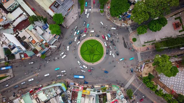 Aerial top-down day timelapse of busy roundabout in Ho Chi Minh City, Vietnam. Fast-moving scooter and car traffic circling star-shaped green island in dynamic Southeast Asian metropolis.