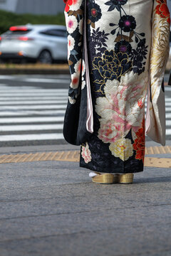 Woman at urban crossing in black and white furisode with ornate floral pattern, wearing accompanying white tabi socks snugly tucked into gold zori to complete traditional fashion coordination. 