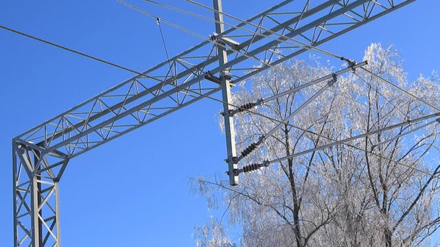 Bottom up view of frozen railway electrification hardware and cables sparkling against blue sky and snowy trees.