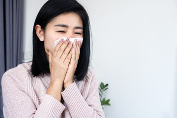 Sick Asian woman blowing nose into tissue, suffering from seasonal flu, common cold, or allergies. Young female feeling unwell with rhinitis symptoms at home. Health care and sickness concept