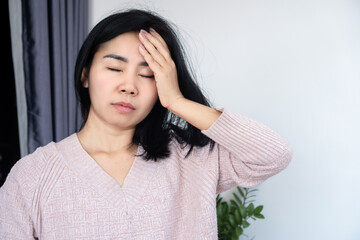 Asian woman suffering from a painful headache, migraine, or vertigo, touching her forehead with eyes closed in discomfort, concept of stress, fatigue, illness, and chronic pain at home indoors