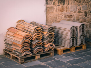 Stacks of weathered terracotta and new gray concrete roof tiles rest on wooden pallets against a rustic stone wall, ready for construction or renovation.