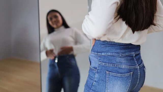 Slim woman looking mirror in modern home interior checking weight loss progress wearing loose jeans at home