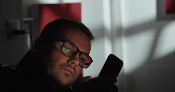 Cinematic close-up of a bored man wearing glasses scrolling on his smartphone indoors, expressing distraction, monotony, and digital overuse.